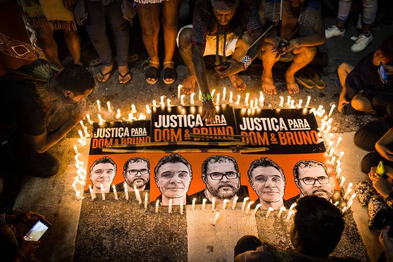 A imagem mostra um grupo de pessoas reunidas em uma manifestação, cercadas por velas acesas. No centro, há cartazes com os dizeres 'JUSTIÇA PARA DOM & BRUNO', acompanhados de ilustrações de rostos. As pessoas estão sentadas no chão, algumas segurando celulares, enquanto outras observam os cartazes e as velas.