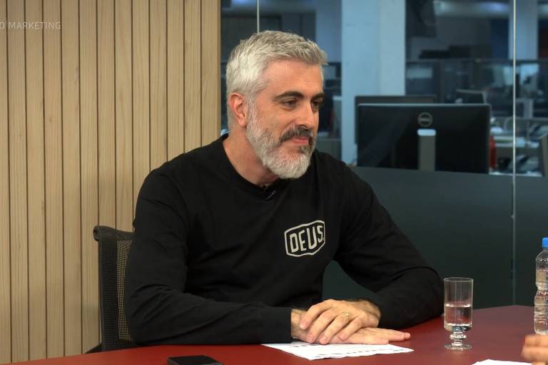 A imagem mostra um homem com cabelo grisalho e barba, sorrindo. Ele está usando uma camiseta preta com a palavra 'DEUS' estampada. Ao fundo, há um ambiente de escritório com mesas e computadores. No canto inferior esquerdo, está escrito 'ARENA DO MARKETING' e 'Burger King'.