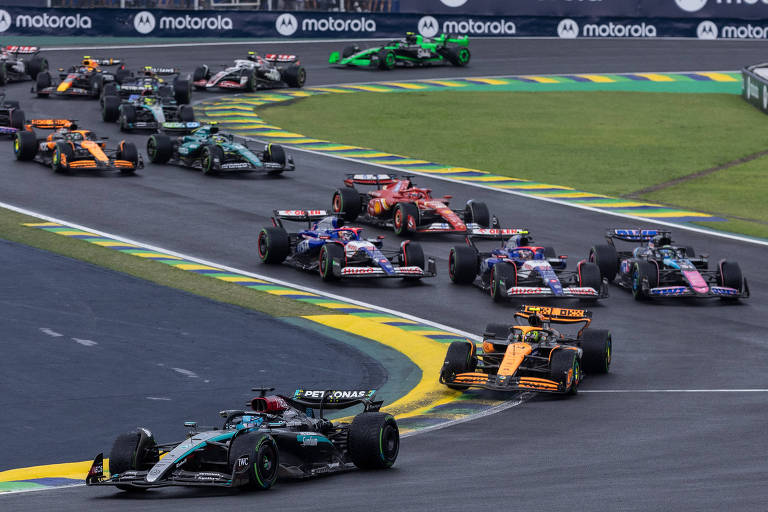 A imagem mostra uma cena de uma corrida de Fórmula 1, com vários carros de corrida em movimento em uma pista. Os carros estão em diferentes posições, alguns em curvas e outros em linha reta. O ambiente é de um circuito, com bandeiras e publicidade ao fundo.