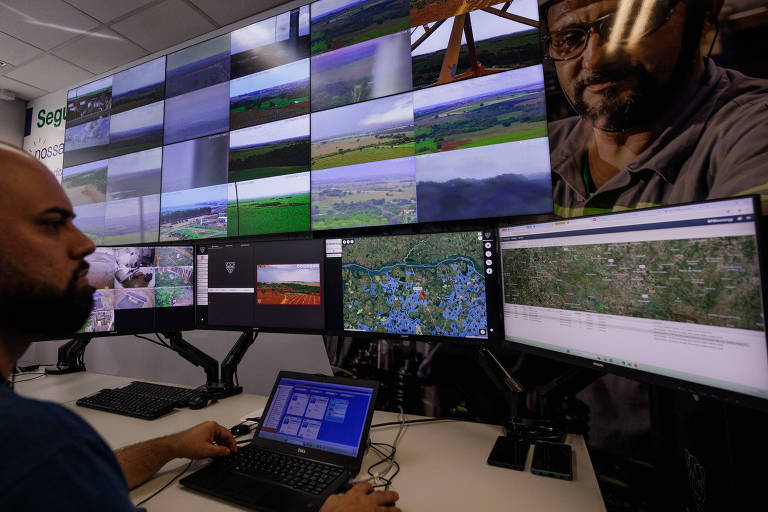 A imagem mostra um centro de monitoramento com várias telas exibindo imagens de câmeras de segurança e um mapa. Um homem está sentado em frente a um computador portátil， com a mão sobre o teclado. As telas na parede mostram diferentes paisagens e áreas monitoradas， enquanto outras telas exibem um mapa detalhado e informações adicionais.