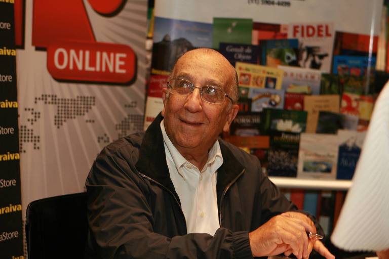 Um homem idoso, usando óculos e uma jaqueta escura, está sentado em uma mesa. Ele parece estar sorrindo ou rindo, com uma expressão amigável. Ao fundo, há uma parede com diversos livros e materiais promocionais, incluindo um banner que diz 'ONLINE'.