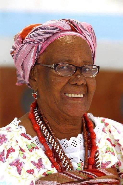 A imagem mostra uma mulher idosa sorrindo, usando óculos e um lenço colorido na cabeça. Ela está vestida com uma blusa branca decorada com flores e colares ao redor do pescoço. O fundo é desfocado, mas parece ser um ambiente interno.