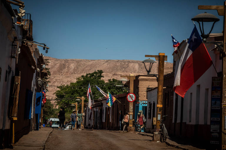 A imagem mostra uma rua em uma cidade chilena, com casas de paredes claras e telhados de barro. Várias bandeiras, incluindo a do Chile, estão penduradas ao longo da rua. Algumas pessoas estão caminhando, e há uma bicicleta encostada em um dos postes. Ao fundo, é possível ver uma montanha árida sob um céu azul claro.