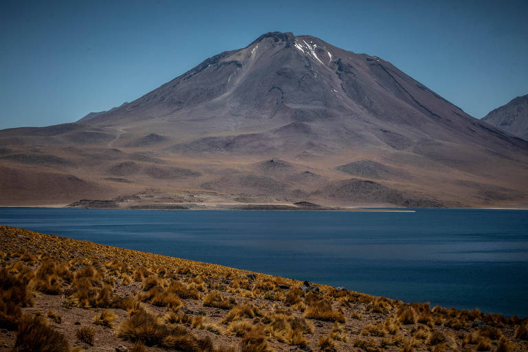 A imagem mostra uma grande montanha com um pico coberto de neve ao fundo， cercada por uma paisagem árida. Na frente， há um lago de água azul， refletindo o céu claro. A vegetação rasteira é visível na margem do lago， contrastando com a terra seca ao redor.