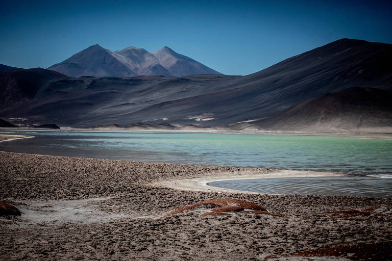 A imagem mostra uma paisagem natural com um lago de águas claras em primeiro plano， cercado por uma área de pedras e areia. Ao fundo， há montanhas com picos visíveis， algumas com uma leve cobertura de neve. O céu está limpo e azul， criando um contraste com as montanhas escuras.