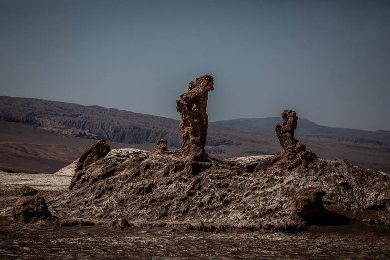 A imagem mostra formações rochosas em um deserto árido, com estruturas irregulares e altas se destacando contra um céu claro. Ao fundo, há montanhas suaves que se estendem até o horizonte.