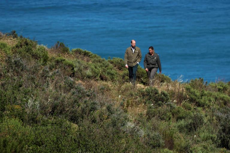 Duas pessoas caminhando em uma área verdejante próxima ao mar. O cenário apresenta um terreno com vegetação rasteira e um fundo de água azul, sugerindo um ambiente costeiro. As pessoas estão vestidas com roupas casuais e parecem estar em uma conversa.