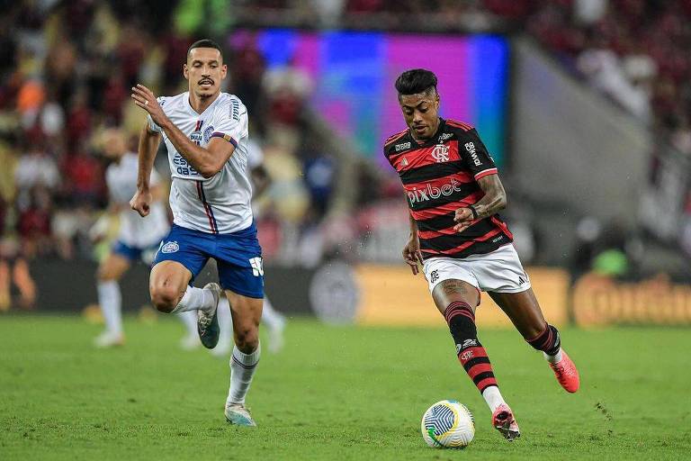 Jogador negro com camisa rubro-negra conduz a bola e é marcado por jogador de camisa brnaca