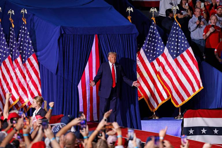 Trump caminha, de braços abertos, sobre uma passarela com sete bandeiras dos Estados Unidos hasteadas a seu redor. Em torno da passarela, apoiadores o filmam e fotografam com celulares. 