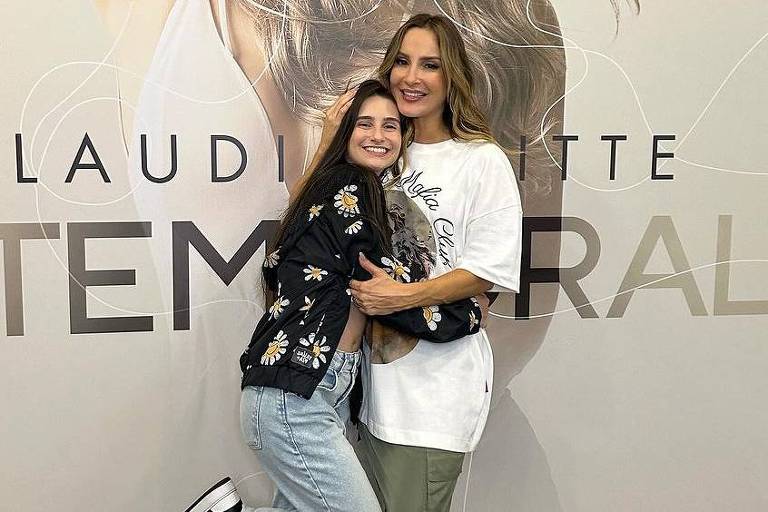 Duas mulheres posam juntas em frente a um grande painel com a imagem de Claudia Leitte. A mulher à esquerda usa uma jaqueta preta com flores, calça jeans e tênis. A mulher à direita veste uma camiseta branca e calças verdes, sorrindo enquanto abraça a outra. O fundo é um painel claro com o nome 'CLAUDIA LEITTE' e a palavra 'IMTEMPORAL' em destaque.