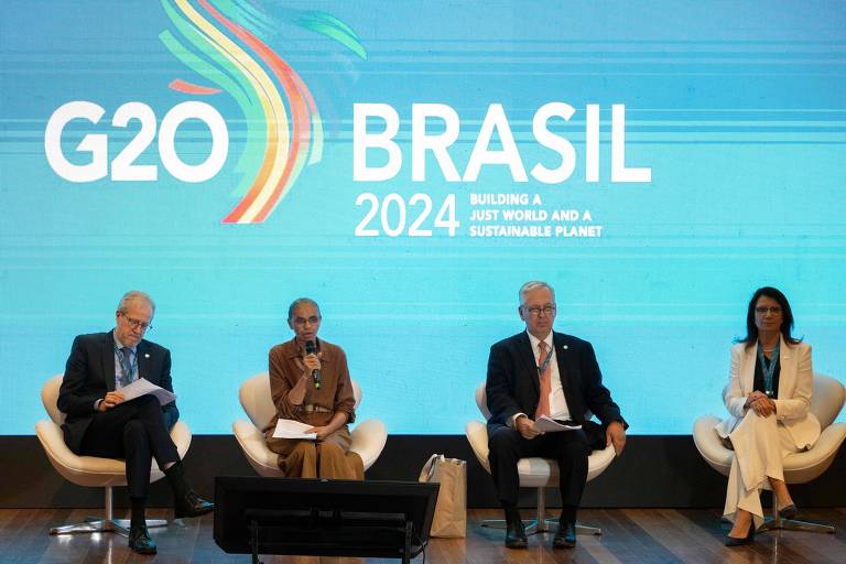 A imagem mostra quatro pessoas sentadas em um painel durante um evento do G20 Brasil 2024. Ao fundo, há um grande banner com as palavras 'G20 BRASIL 2024' e um design gráfico colorido. As pessoas estão vestidas formalmente, com algumas segurando papéis e outras com as mãos no colo.