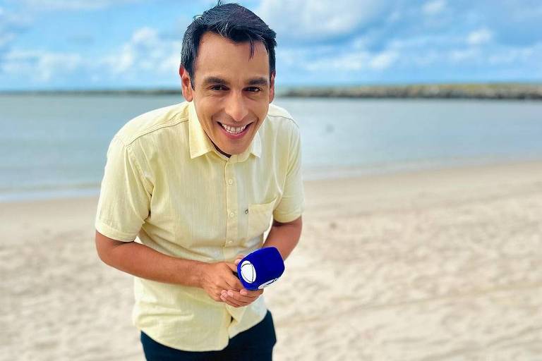 Um homem sorridente está posando na praia, segurando um microfone azul. Ele usa uma camisa amarela e calças escuras, com sapatos marrons. Ao fundo, vê-se o mar e um céu parcialmente nublado.