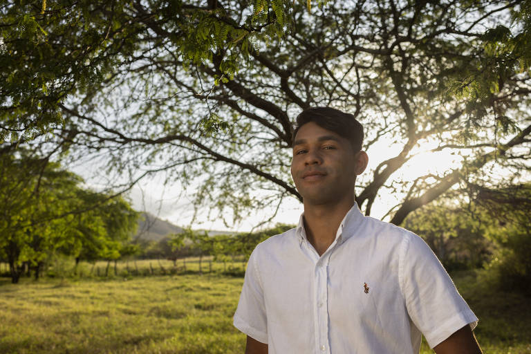 Um jovem está posando em um campo aberto, com uma árvore grande ao fundo. Ele usa uma camisa branca e tem um leve sorriso no rosto. A luz do sol brilha por trás dele, criando um efeito de silhueta. O ambiente é verde e natural, com grama e árvores ao redor.