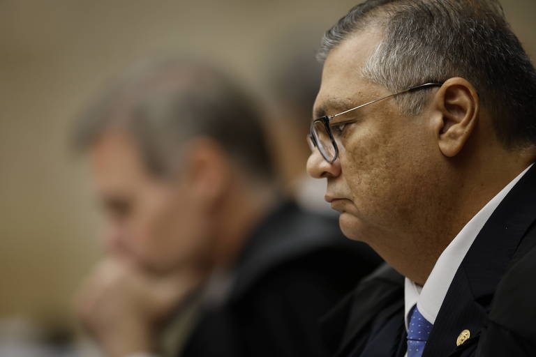 A imagem mostra um grupo de homens em uma reunião. Em primeiro plano, um homem de óculos e cabelo curto está olhando para baixo, com uma expressão séria. Ao fundo, outro homem está parcialmente visível, também com uma expressão concentrada. O ambiente parece ser um espaço formal, possivelmente uma sala de reuniões.