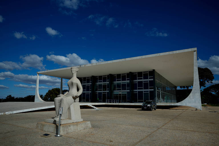 A imagem mostra um edifício moderno com uma fachada de vidro e um telhado em forma de asa. Em primeiro plano, há uma escultura de uma figura humana em pedra. O céu está limpo com algumas nuvens brancas