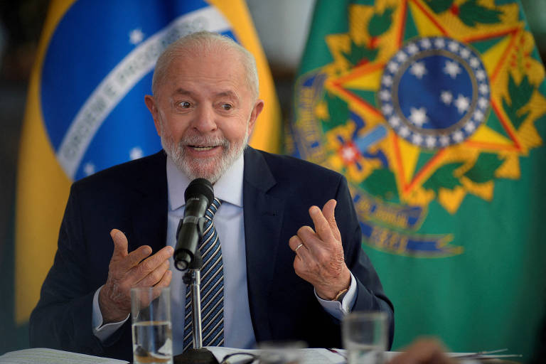 homem branco de cabelo e barba branca gesticula com as duas mãos. Ele usa terno e gravata. Atrás, há bandeira e brasão do Brasil