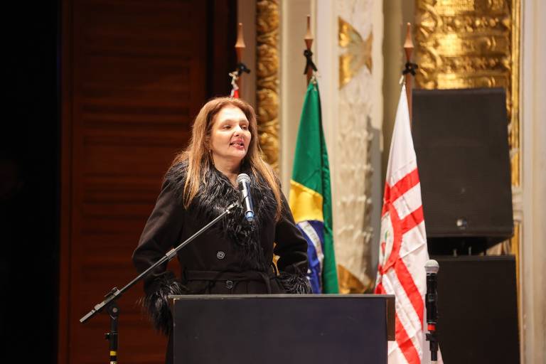 Uma mulher em pé atrás de um púlpito, falando em um evento. Ela usa uma roupa escura e tem cabelo longo e solto. Ao fundo, há bandeiras do Brasil e de São Paulo, além de um painel decorativo. O ambiente parece ser um auditório ou sala de conferências.