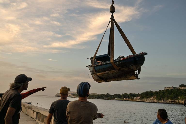 A imagem mostra um barco sendo içado por um guindaste em um porto. Quatro homens estão observando o processo, com um deles apontando para o barco. O céu está parcialmente nublado, com tons de azul e laranja ao entardecer. Ao fundo, é possível ver a água e algumas construções na margem.