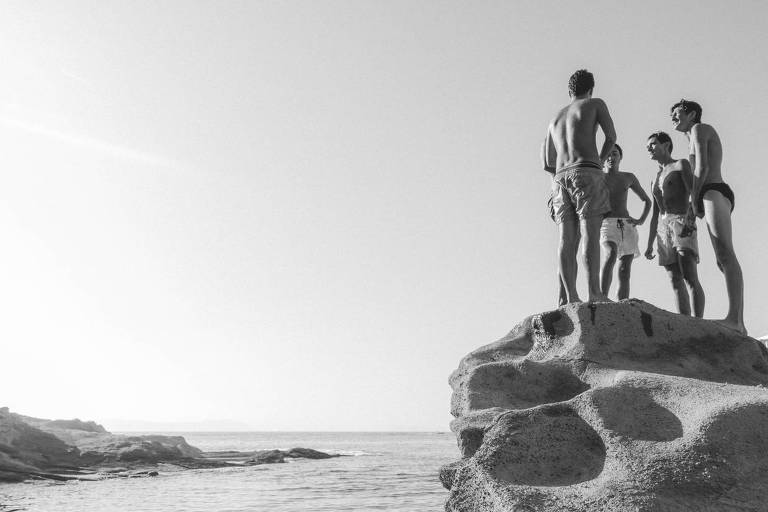 Cinco jovens estão em uma rocha à beira-mar, conversando. Eles estão sem camisa e vestindo trajes de banho. O cenário é de uma praia com água calma e rochas ao fundo, sob um céu claro. A imagem é em preto e branco.