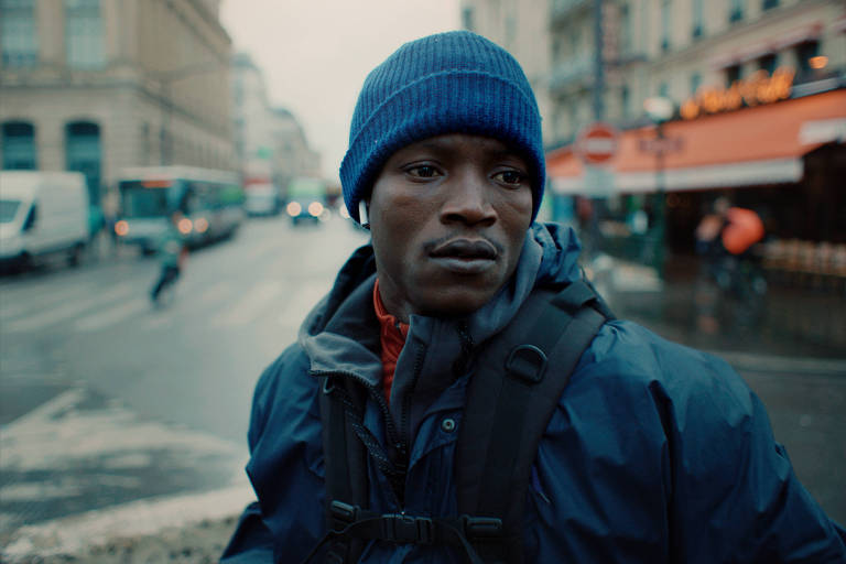 A imagem mostra um homem com um boné azul e fones de ouvido, olhando para o lado em uma rua urbana. Ele está usando uma jaqueta escura e uma mochila. Ao fundo, há veículos e edifícios, com um clima nublado e chuvoso.