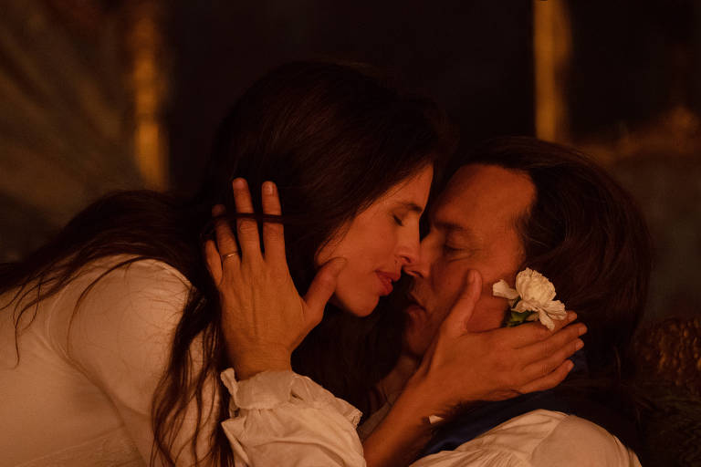 A imagem mostra um casal em um momento íntimo, com os rostos quase se tocando. A mulher tem cabelos longos e escuros, e está vestindo uma blusa branca. O homem, com cabelos longos e soltos, usa uma camisa clara e um lenço azul. Ele segura uma flor branca perto do rosto. O fundo é difuso, sugerindo um ambiente acolhedor e romântico.