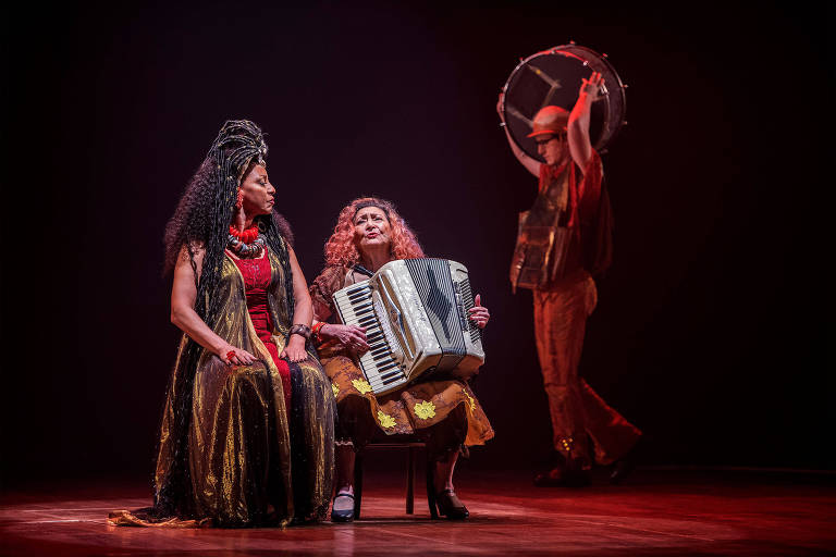 A imagem mostra duas mulheres sentadas em um palco. A mulher à esquerda tem cabelo crespo e usa um vestido longo e brilhante, com colares vermelhos. A mulher à direita, com cabelo cacheado e vestido amarelo com flores, toca um acordeão. Ao fundo, um homem vestido com roupas metálicas e um chapéu toca um tambor. A iluminação é suave, criando um ambiente dramático.