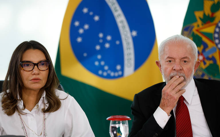 Foto mostra primeira-dama Janja e o presidente Lula. Ela está do lado esquerdo da imagem e ele do lado direito. O casal tem pele clara. Janja tem cabelos castanhos na altura dos ombros e usa óculos com bordas grossas. Ela está séria e veste um paletó branco por cima de uma camisa branca adornada por um colar dourado. Seu esposo, veste um terno preto com camisa branca e uma gravata vermelha. Ele tem barba e cabelos grisalhos com sinais de calvície. Lula está com a mão direita na frente dos lábios. Atrás do casal, que está sentado, é possível observar uma bandeira do Brasil e outra da república.