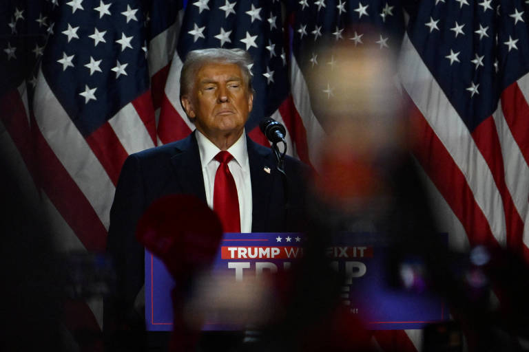 A imagem mostra um homem em pé em um palco, com bandeiras dos Estados Unidos ao fundo. Ele está usando um terno escuro e uma gravata vermelha, com uma expressão séria. À sua frente, há um cartaz com as palavras 'TRUMP' e 'WILL FIX IT'. A imagem está ligeiramente desfocada na parte inferior, com algumas silhuetas de pessoas visíveis.