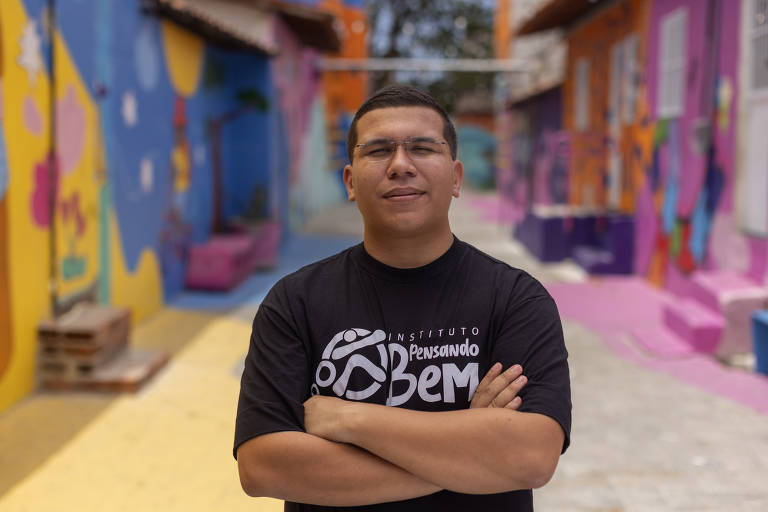 Um jovem está posando em um espaço urbano colorido, com paredes decoradas em tons vibrantes de azul, rosa e laranja. Ele usa uma camiseta preta com o logo do 'Instituto Pensando Bem'. O jovem tem os braços cruzados e um leve sorriso, enquanto o fundo apresenta um caminho pavimentado e áreas com bancos e murais artísticos.