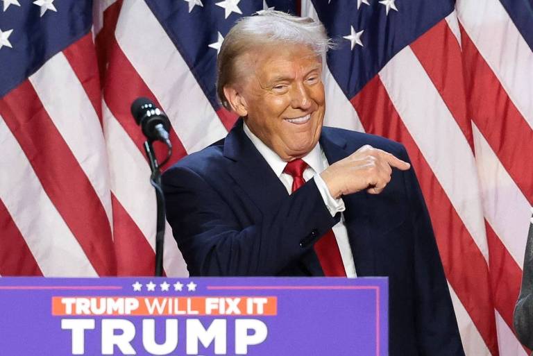 A imagem mostra Donald Trump sorrindo e apontando para uma mulher ao seu lado, que está aplaudindo. Ao fundo, há várias bandeiras dos Estados Unidos dispostas em fileiras. Na parte inferior da imagem, há um banner com o texto 'TRUMP WILL FIX IT VANCE 2024'.
