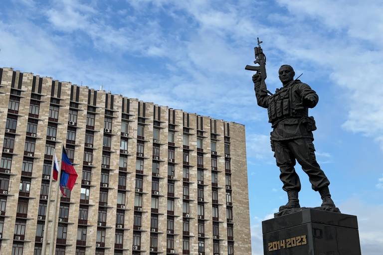 Estátua com soldado empunhando arma com a inscrição 2014-2023 abaixo, em frente a prédio cor de creme com bandeiras