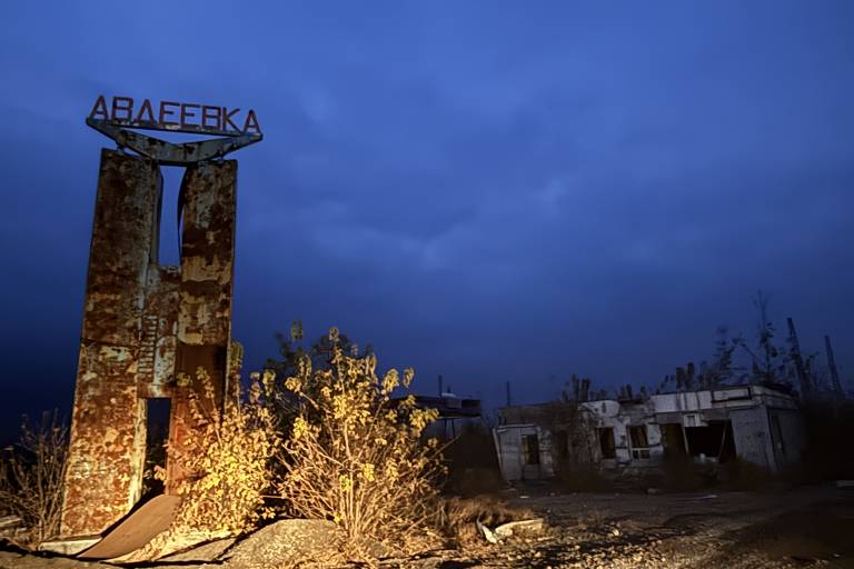 Monumento com Avdiivka escrito em cirílico em cima. A foto é noturna e a estrutura está iluminada por faróis. Ruínas ao fundo