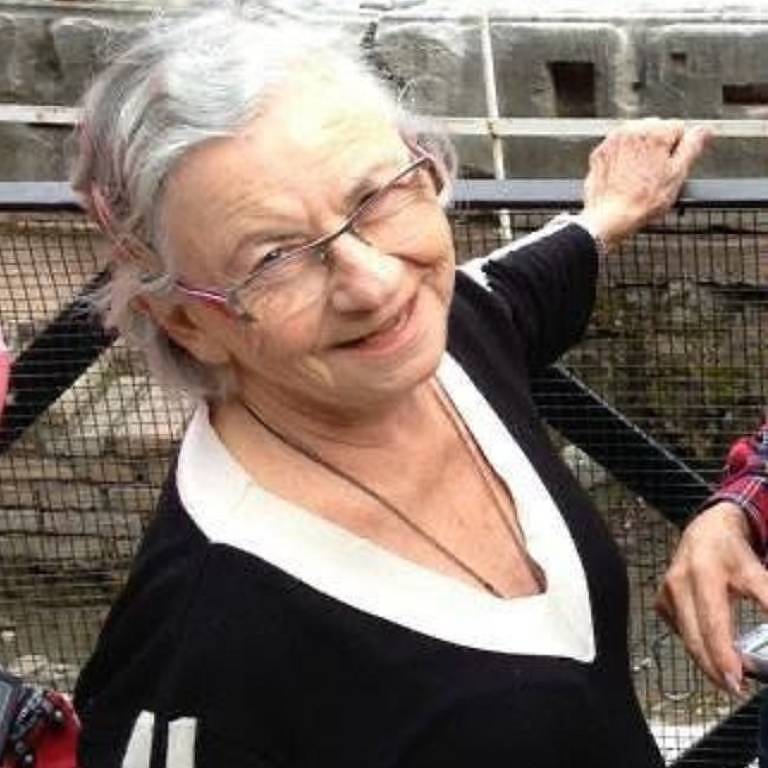 Mulher idosa com cabelo grisalho e óculos, sorrindo enquanto se apoia em uma grade. Ela está vestindo uma blusa preta com detalhes brancos e parece estar em um local histórico, com paredes de pedra ao fundo