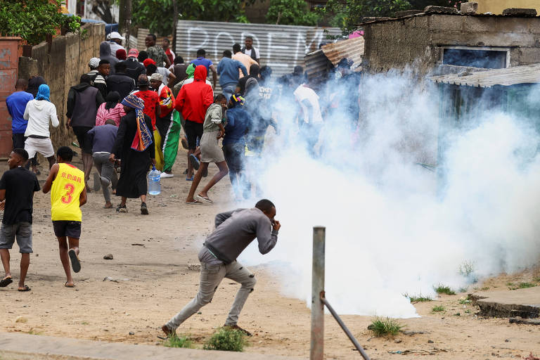 A imagem mostra um grupo de pessoas em uma área urbana, algumas delas correndo em direção a uma nuvem de fumaça. Um homem em primeiro plano está se agachando e cobrindo o rosto, enquanto outros indivíduos estão dispersos ao fundo. A cena parece ser de um protesto ou confronto, com fumaça visível no ar e edifícios simples ao fundo.