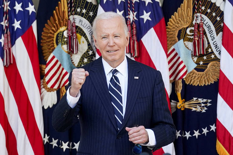 Um homem de cabelo grisalho, vestindo um terno escuro e uma gravata listrada, está posando com o punho cerrado em um gesto de determinação. Ao fundo, há várias bandeiras dos Estados Unidos e emblemas presidenciais.