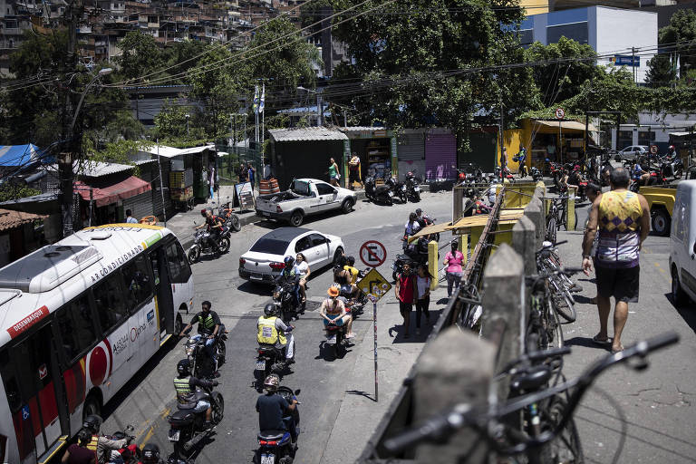 A imagem mostra uma área urbana movimentada, com um ônibus, carros e várias motocicletas em uma rua. À direita, uma pessoa caminha ao longo de uma calçada. Ao fundo, há barracos e estruturas comerciais, além de vegetação. O céu está claro e a cena parece ser em um dia ensolarado.