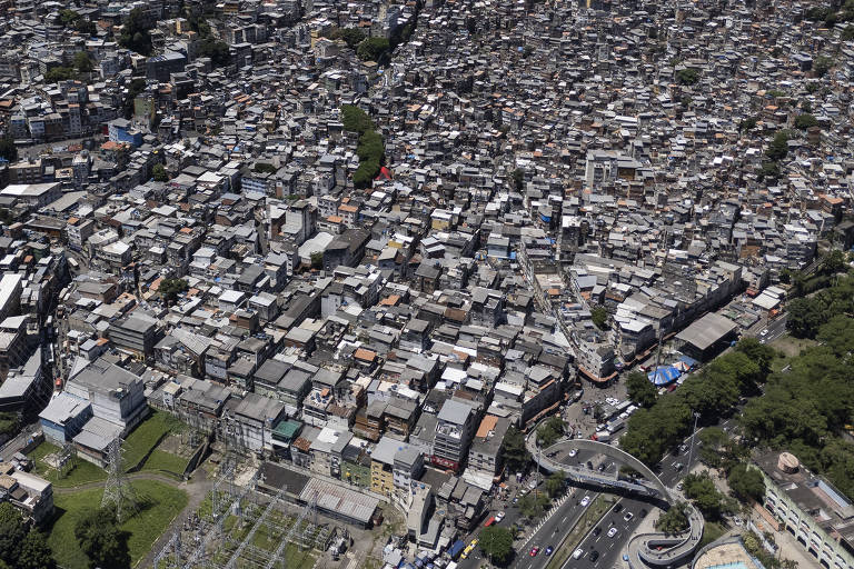 Imagem aérea de uma área urbana densamente povoada, com muitas casas e edifícios próximos uns dos outros. A disposição das construções é irregular, com telhados de diferentes cores e tamanhos. Na parte inferior da imagem, há uma estrada com veículos e uma área verde ao lado. A imagem mostra uma grande concentração de habitações, típica de comunidades urbanas.