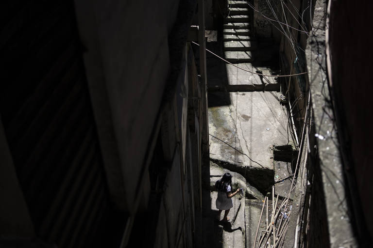 Rua estreita com escadarias, fios expostos e grande sombra das casas sobre a rua. Uma menina parece andar por ali