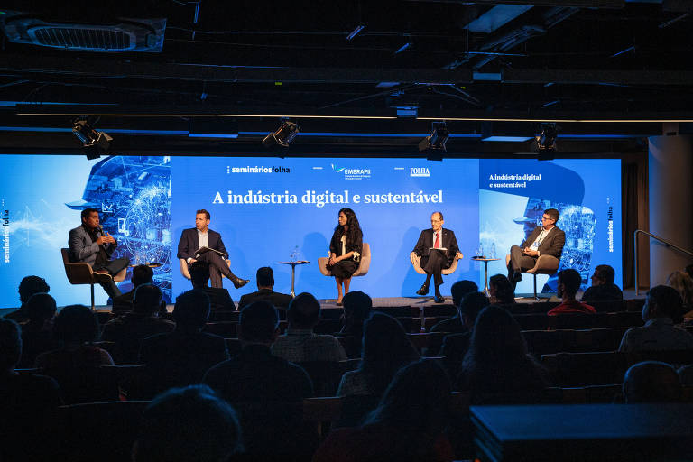 A imagem mostra um painel de discussão com cinco pessoas sentadas em cadeiras em um palco. Ao fundo, há um telão azul com a frase 'A indústria digital e sustentável'. O ambiente parece ser um auditório, com uma plateia visível em frente ao painel. Os participantes estão vestidos formalmente e parecem estar envolvidos na discussão.