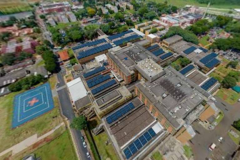 A imagem mostra uma vista aérea de um complexo educacional, com vários edifícios dispostos em um layout organizado. Os telhados dos prédios são cobertos com painéis solares. Na parte inferior esquerda, há um campo esportivo com uma quadra azul. Ao fundo, pode-se ver áreas verdes e outros edifícios ao redor.