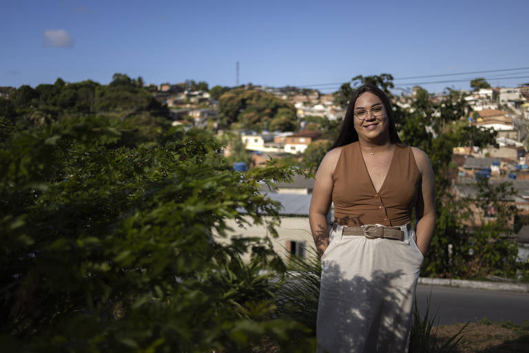 Uma mulher está em pé em um ambiente ao ar livre, com uma paisagem urbana ao fundo. Ela usa uma blusa marrom e uma saia clara, com as mãos nos bolsos. O cenário inclui casas em uma colina e um céu azul com algumas nuvens. A vegetação verde está em primeiro plano, em destaque.