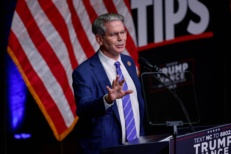 NA foto， um homem branco grisalho de terno e gravatas azuis discursa em um palanque com o nome de Trump， e uma bandeira dos Estados Unidos ao fundo