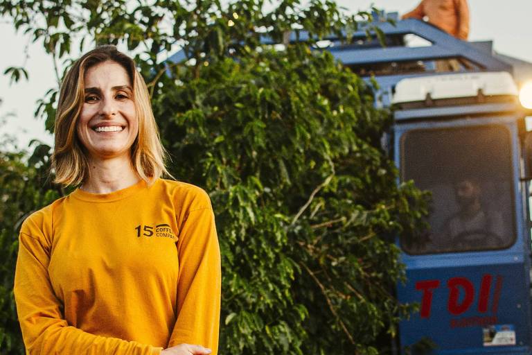 A imagem mostra uma mulher sorridente em uma plantação de café， vestindo uma camiseta de manga longa amarela com o logo 039;15 Coffee Company039;. Ela está de pé， com os braços cruzados， e segura um par de óculos de sol. Ao fundo， há uma máquina de colheita de café azul e um homem com chapéu em cima dela. A vegetação ao redor é densa， com folhas verdes.