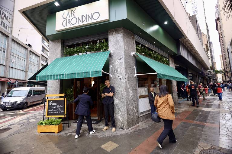 A imagem mostra a fachada de um restaurante chamado 'Café Urânimo'. O local possui toldos verdes e uma placa de madeira na entrada. Há pessoas caminhando na calçada e um carro estacionado próximo ao restaurante. O ambiente é urbano, com prédios altos ao fundo.