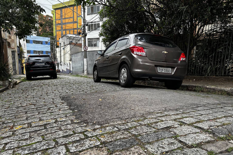 A imagem mostra uma rua de paralelepípedos com dois carros estacionados. À esquerda, um carro preto e à direita, um carro cinza. Ao fundo, há prédios com fachadas em cores variadas, incluindo um prédio amarelo e azul. A vegetação está presente nas laterais da rua, e há fios elétricos visíveis.