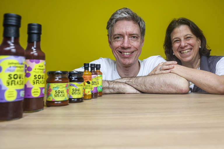 A imagem mostra um homem e uma mulher sorrindo, posando em frente a uma mesa com vários frascos de produtos alimentícios. O fundo é amarelo e os frascos incluem molhos e conservas, todos com rótulos coloridos. O homem está com os braços cruzados sobre a mesa, enquanto a mulher está ao seu lado, apoiando-se levemente.