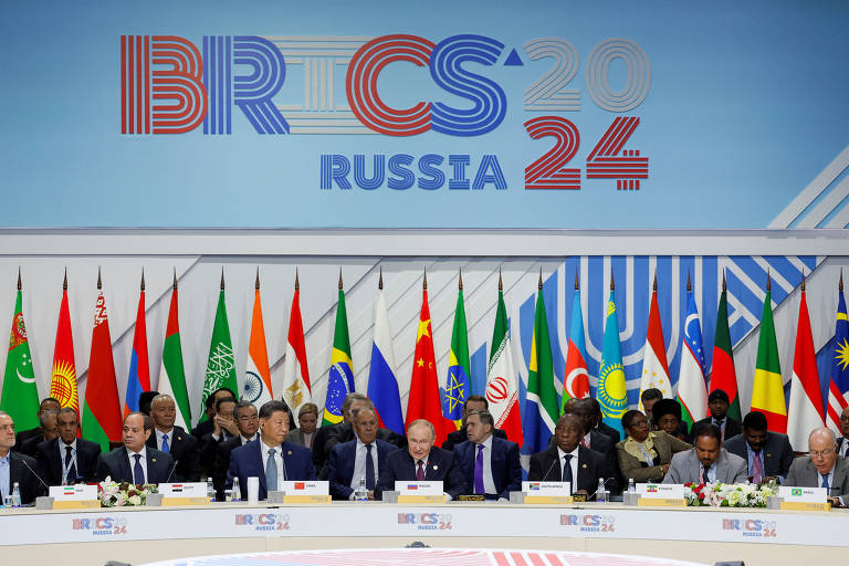 A imagem mostra uma mesa de conferência com vários líderes sentados, em frente a uma parede decorada com o logotipo 'BRICS 24' e a palavra 'Rússia'. Atrás deles, há uma exibição de bandeiras de diferentes países, representando os membros do BRICS.
