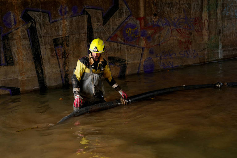 Um trabalhador está em uma área alagada, usando um capacete amarelo e roupas de proteção. Ele está segurando um tubo preto enquanto a água reflete ao seu redor. Ao fundo, há uma parede com grafites coloridos.