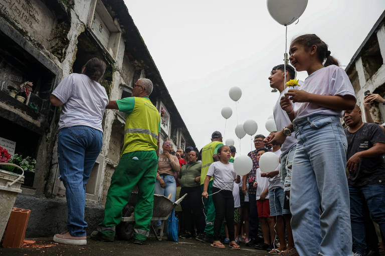 A imagem mostra um grupo de pessoas em um cemitério, algumas delas segurando balões brancos. Um homem vestido de verde, possivelmente um funcionário do cemitério, está ao lado de um túmulo, enquanto outras pessoas observam. A atmosfera parece ser de homenagem ou celebração, com flores e um ambiente nublado ao fundo.