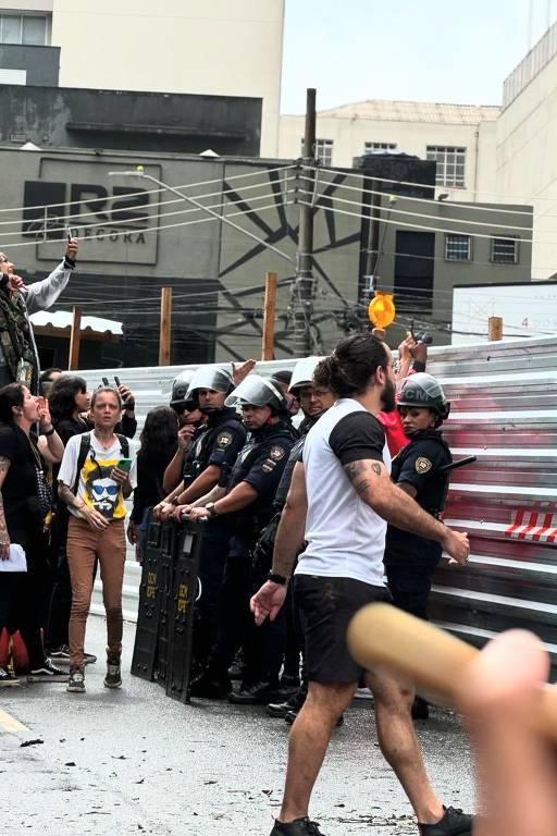 A imagem mostra um confronto entre manifestantes e policiais em um ambiente urbano. No primeiro plano, há um grupo de manifestantes, alguns com expressões de protesto, enquanto outros estão em uma posição mais calma. Os policiais, em uniformes e capacetes, estão alinhados em uma barreira, segurando escudos. Ao fundo, há edifícios e fios expostos, além de uma faixa de segurança. O clima parece tenso, com a presença de objetos de barricada.